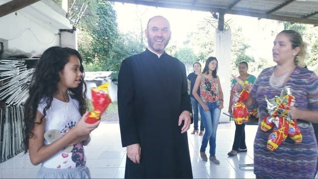 Menina com o presente recebido, ao lado do sacerdote e de uma voluntária