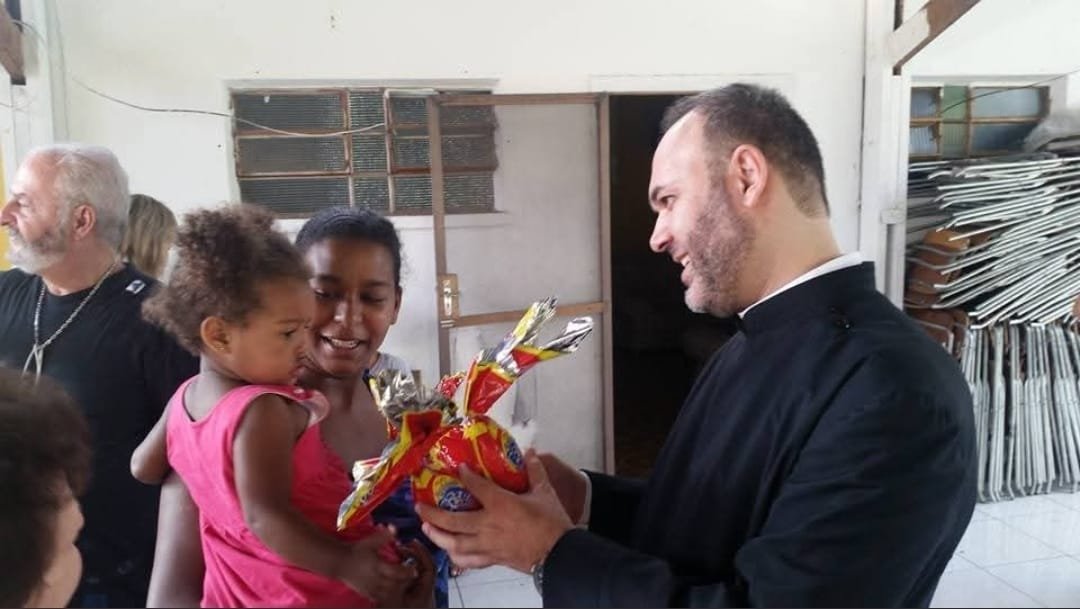 Sacerdote entrega presente a uma criança no colo da mãe